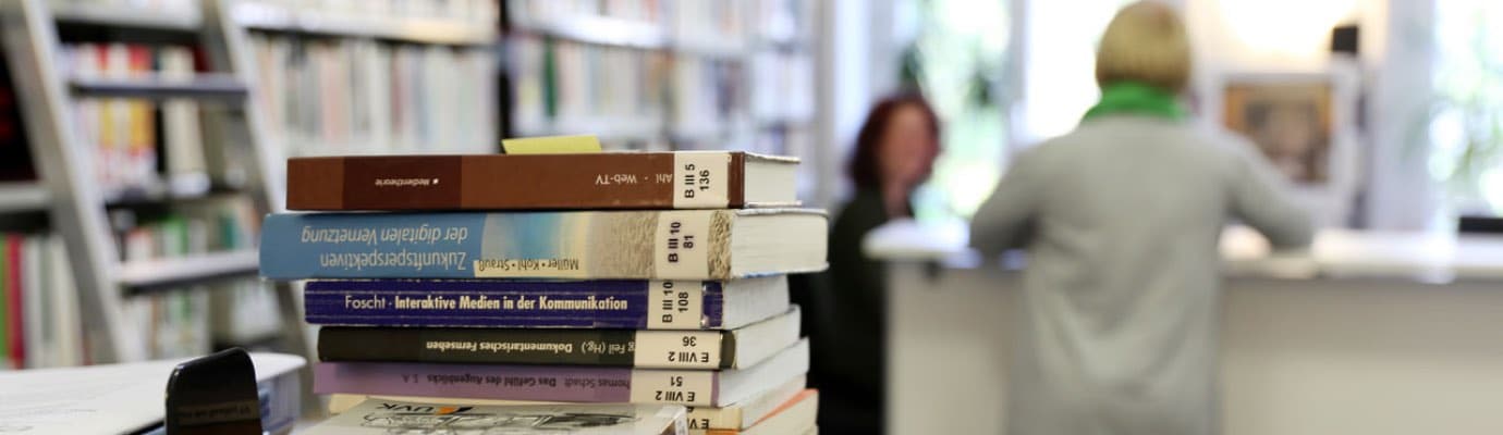 Blick in eine Bibliothek mit einem Bücherstapel im Vordergrund