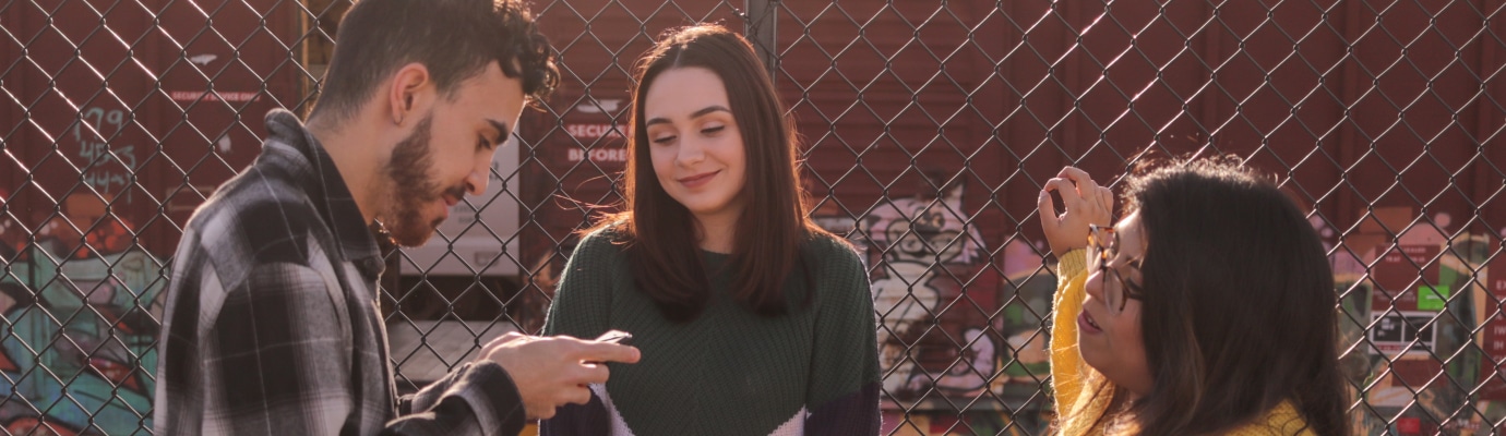 Three people leaning against a wire mesh fence, one is looking at his smartphone