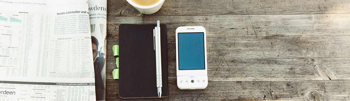 Zeitung, Notizbuch mit Stift, Mobiltelefon, Kaffeebecher und Sonnenbrill auf einem Holztisch.