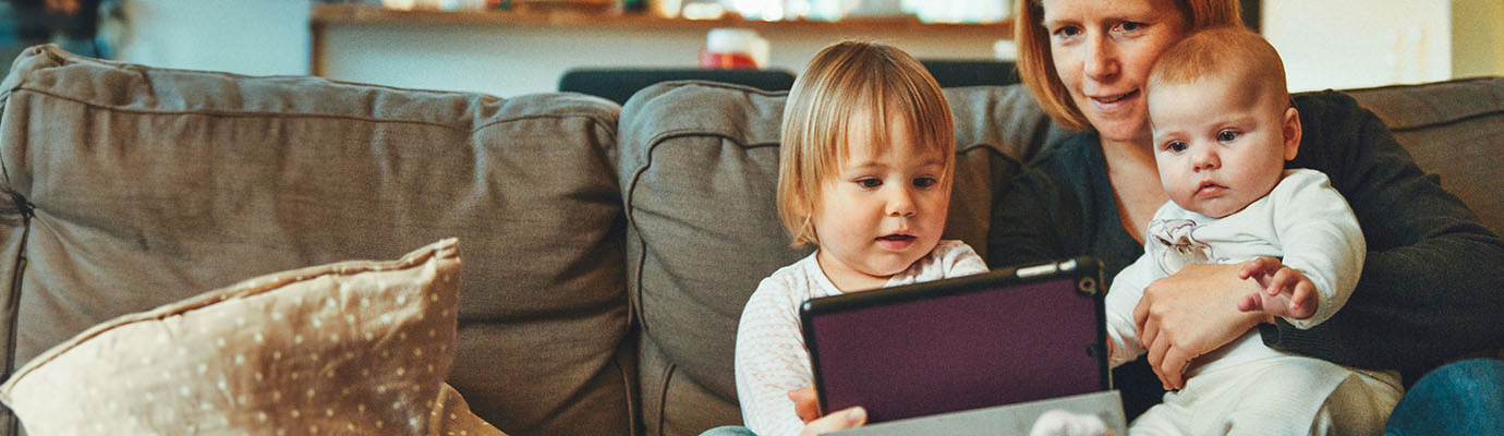 Auf der Couch sitzende Mutter guckt mit Baby und Kleinkind auf ein Tablett