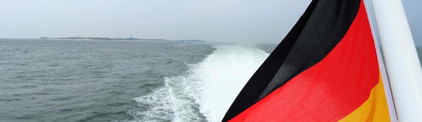 Deutschlandflagge weht an Schiffsheck. Dahinter Fahrwasser und weites Meer