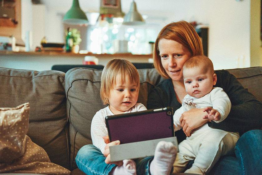 Auf der Couch sitzende Mutter guckt mit Baby und Kleinkind auf ein Tablett