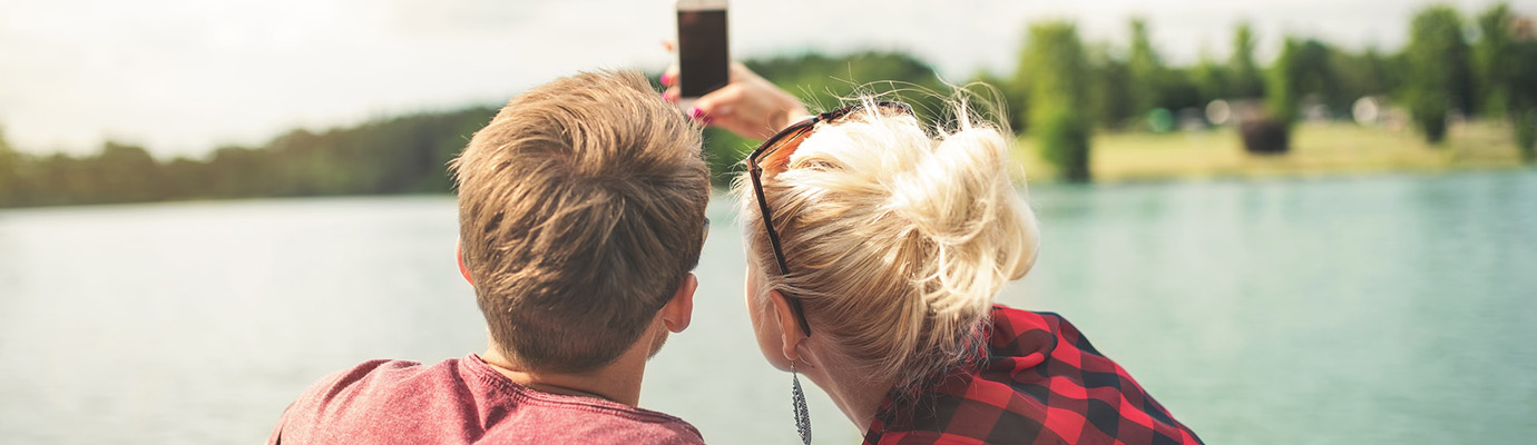 Von hinten zu sehen: Blondes junges Paar macht Selfie am See