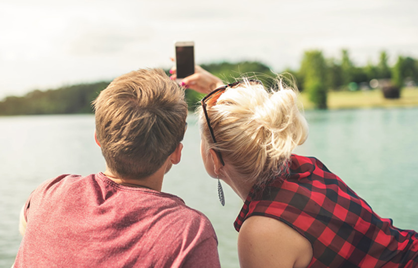 Von hinten zu sehen: Blondes junges Paar macht Selfie am See