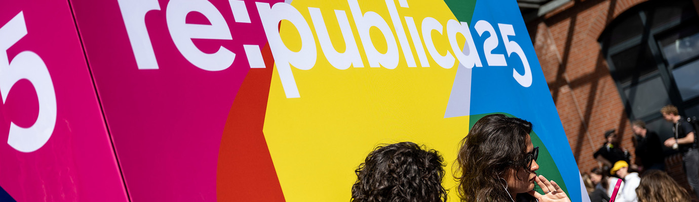 zwei junge Frauen sitzen auf ihr Handy blickend vor einem großen rot-gelb-grün-blauen Banner mit RePublica25-Logo