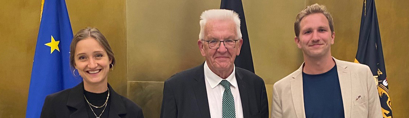 Leonie Alatassi, Ministerpräsident Winfried Kretschmann und Kilian Hampel vor goldener Wand mit Europaflagge