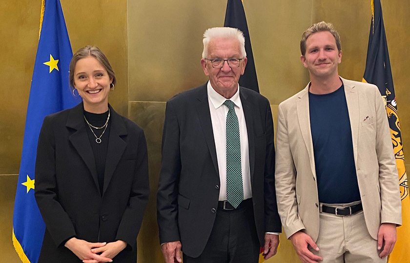 Leonie Alatassi, Ministerpräsident Winfried Kretschmann und Kilian Hampel vor goldener Wand mit Europaflagge